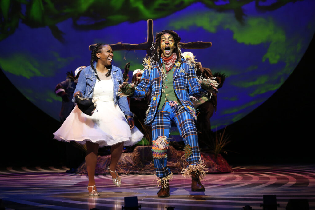 Dorothy laughs as the Scarecrow sings and dances. She wears a white dress, a denim jacket and silver shoes. The Scarecrow wears a bright blue plaid suit with colorful patches and a green vest.