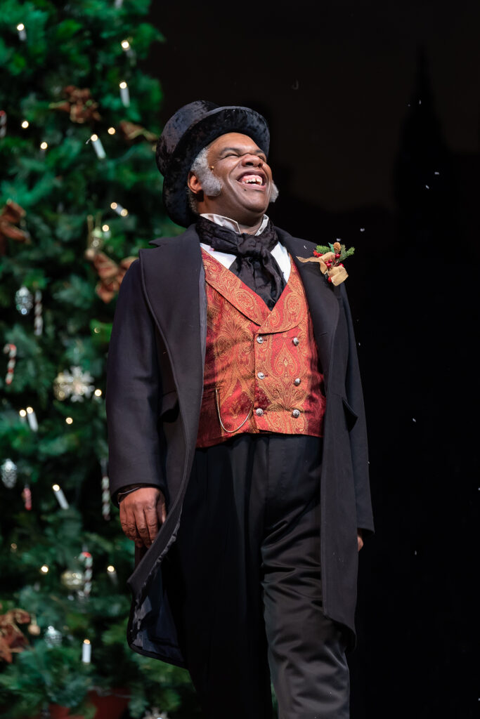 A smiling man in Victorian-style clothes and a top hat stands in front of a Christmas tree.