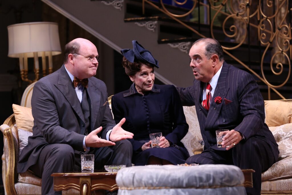 Two older men and an older woman, all in 1950's style outfits, sit on a long sofa and talk animatedly. Two hold drinks in their hands, one has put his drink down on a table in front of them.