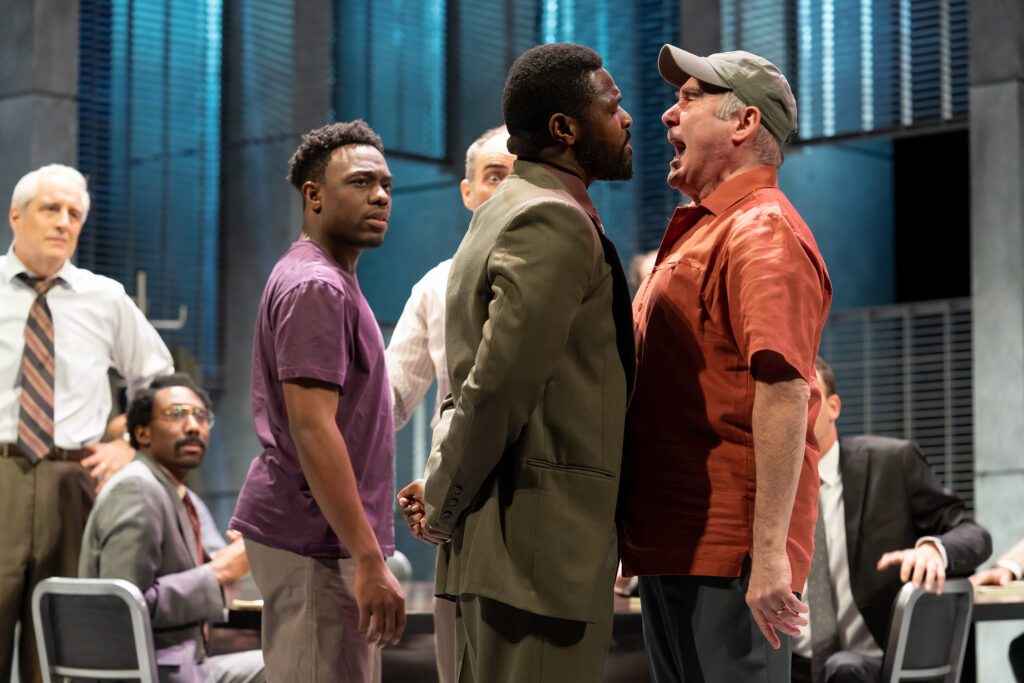 Four men stand in a jury room, where other jurors are seated. A white man in a ballcap (actor Lawrence Redmond) yells in the face of a black man (actor Bueka Uwemedimo) with whom he disagrees.
