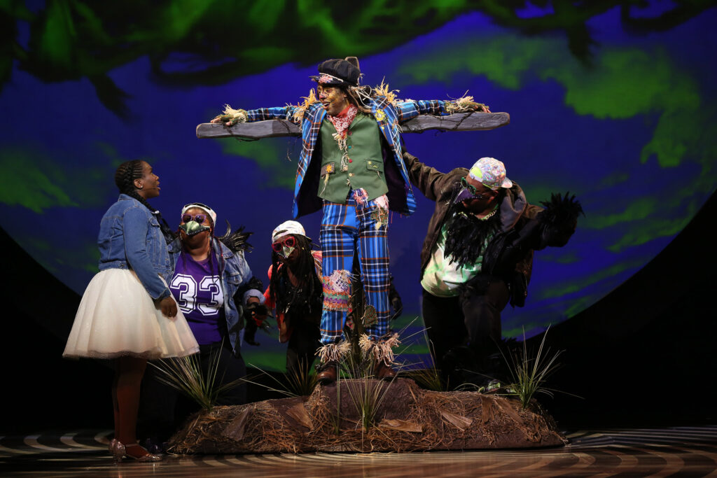 Dorothy looks up at the Scarecrow, who is tied to a post. He is surrounded by crows. She wears a white dress, a denim jacket and silver shoes. The Scarecrow wears a bright blue plaid suit with colorful patches and a green vest. The crows wear backward baseball caps, t-shirts and jackets.