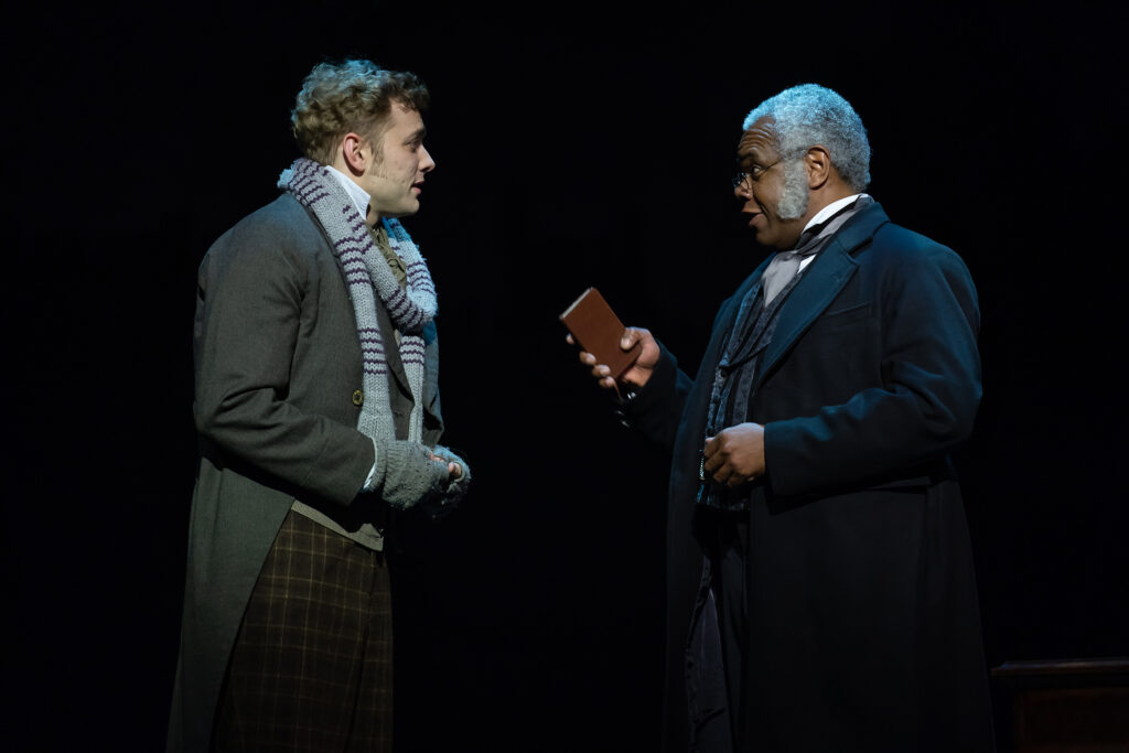 A gray-haired older man dressed in Victorian-clothes holds a small notebook and speaks to a younger man also dressed in Victorian dress.
