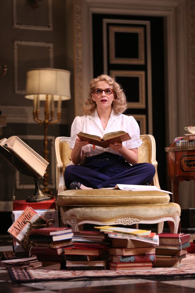 A woman in 1950's clothing sits cross-legged on a fancy chair and stares straight ahead. She holds a large book in her hands, and another pile of books are strewn on the floor in front of her.