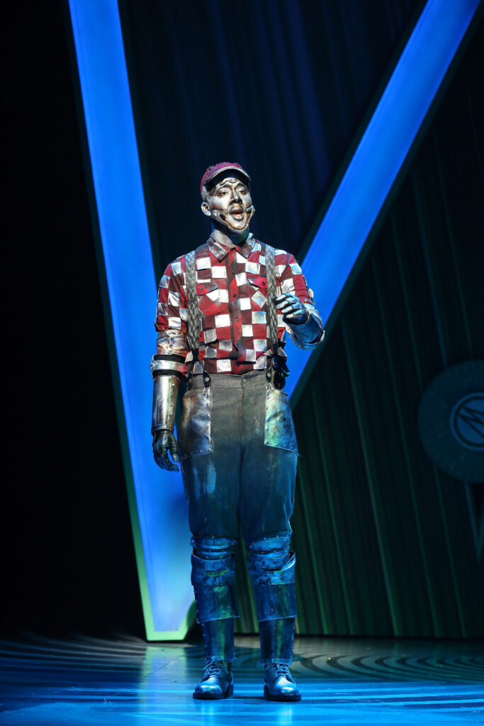 The Tinman sings. He wears a red-and-silver checkered shirt and silver pants.