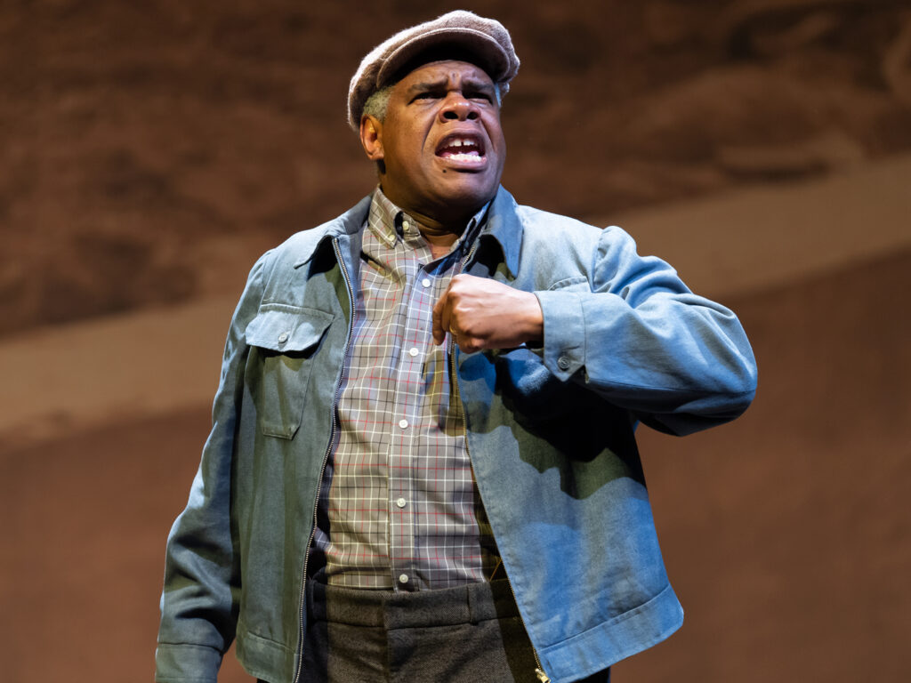 A man shouts into the sky in his backyard. He wears a brown cap, a casual blue jacket over his checkered button-down collared shirt and woolen pants.