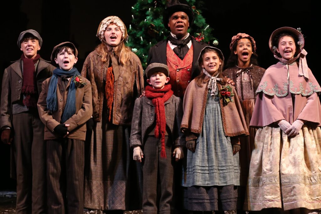 Scrooge and a group of children sing while standing in front of a tall Christmas tree.