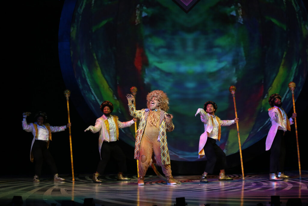At center, the Lion dances and sings. He is surrounded by the yellow brick road, played by four dancers with black-and-gold afros who wear white tailcoats with gold trim, gold sneakers, black pants, white shirts and white vests. They carry tall gold scepters.