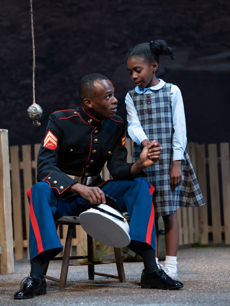 A man in a Marine uniform sits on a stool in his childhood backyard, holding the hand of his six-year-old half-sister as they talk about their father.