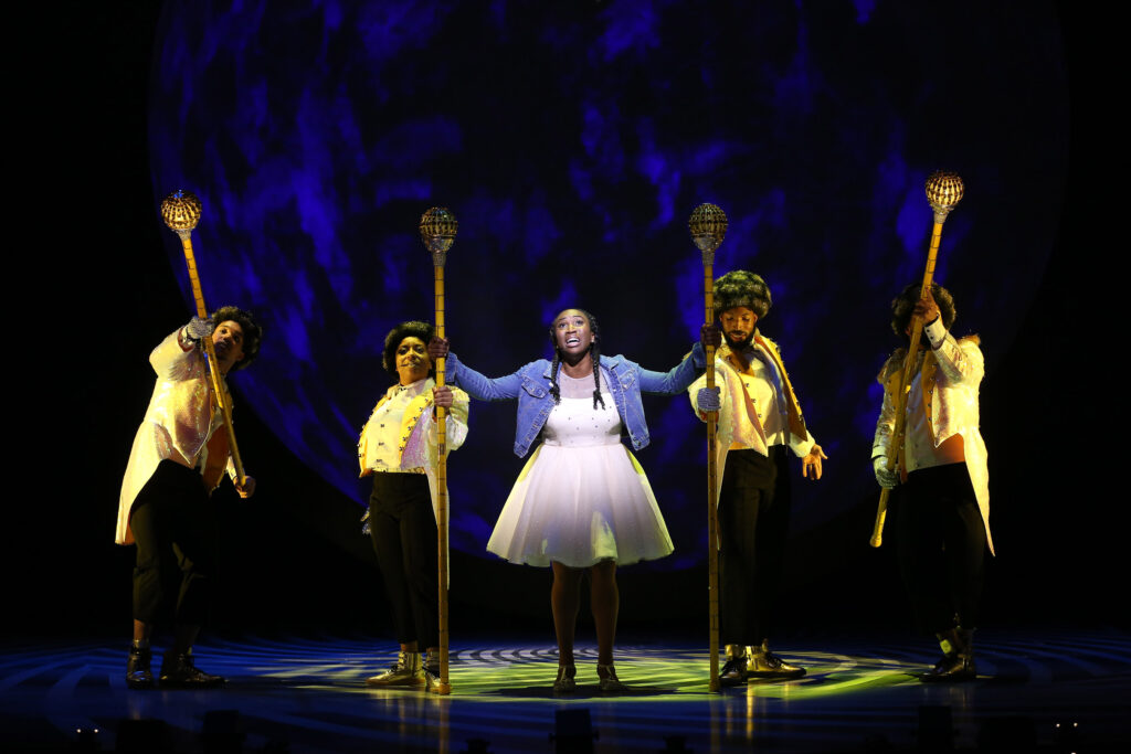 At center, Dorothy sings. She is surrounded by the yellow brick road, played by four dancers with black-and-gold afros who wear white tailcoats with gold trim, gold sneakers, black pants, white shirts and white vests. They carry tall gold scepters.