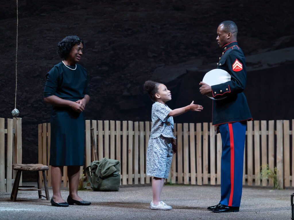 A man in a Marine uniform shakes the hands of his six-year-old half-sister in their backyard. His mother looks on.