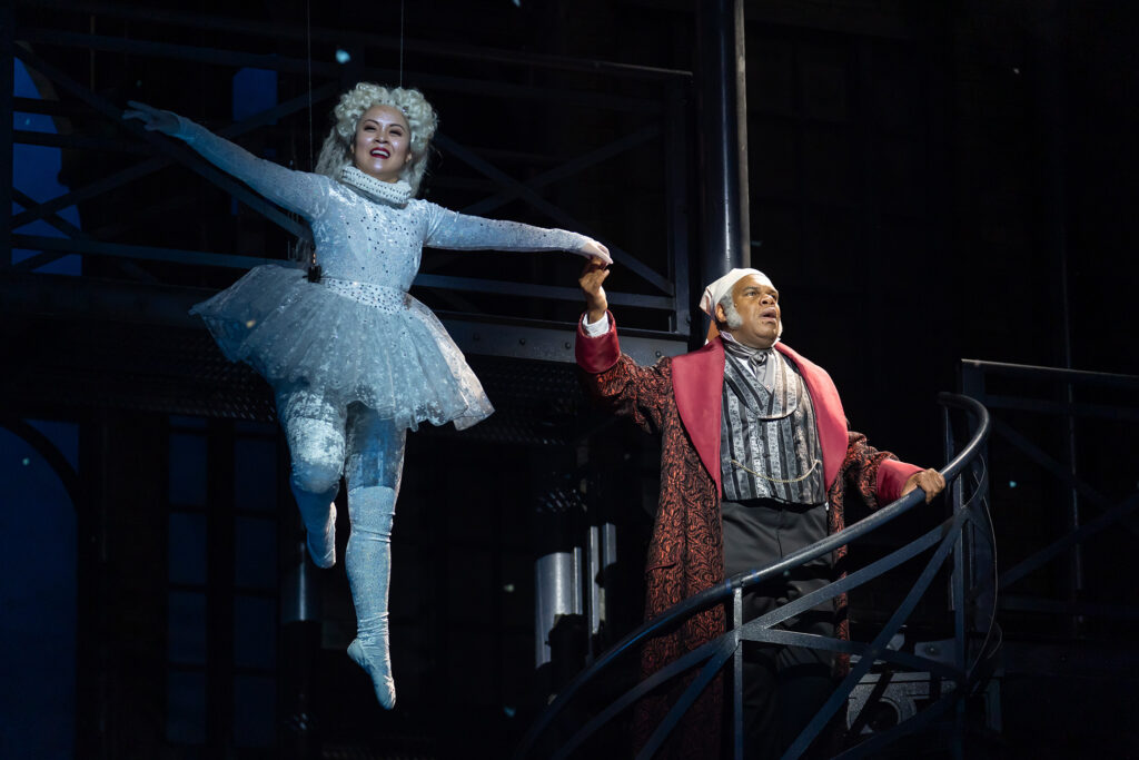 A joyful spirit flies through the air in a sparkling, long-sleeved unitard and matching tutu. She grasps the hand of Ebenezer Scrooge and the two glide upward.