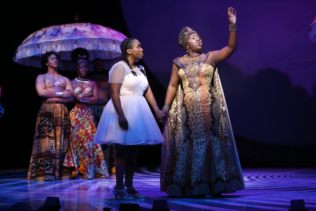 Dorothy holds the hand of Glinda, as Glinda sings. Dorothy wears a white dress and silver shoes. Glinda wears an earth-toned full-length dress, necklace and head-dress inspired by Afro-futurism. In the background stand a shirtless man in a full-length skirt and a woman in a shiny midriff halter shirt and full-length skirt; their clothing is also inspired by Afro-futurism.