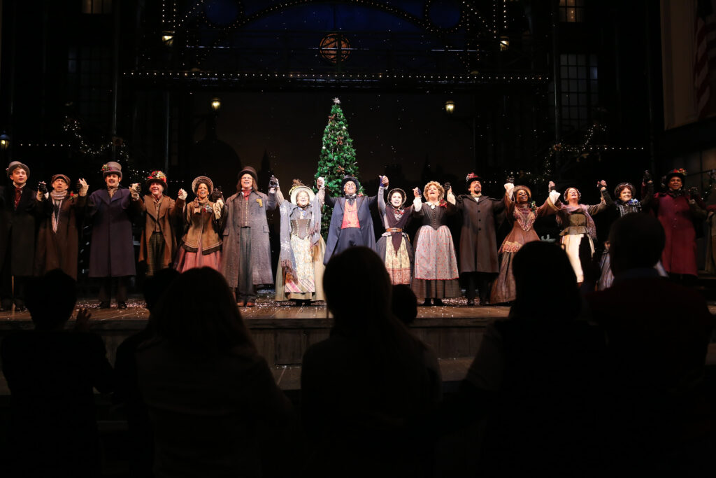 The entire cast stands in front of a giant Christmas Tree and raises their hands to bow. The silhouettes of the audience can be seen in front of them.