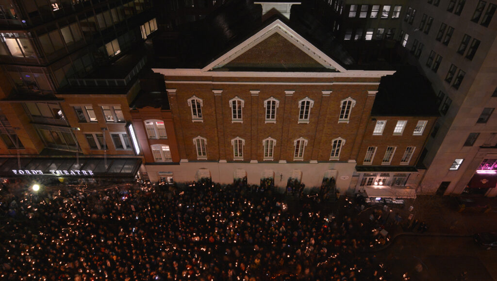 A large crown of people gather in front of Ford's Theatre at night. Most hold lit candles.