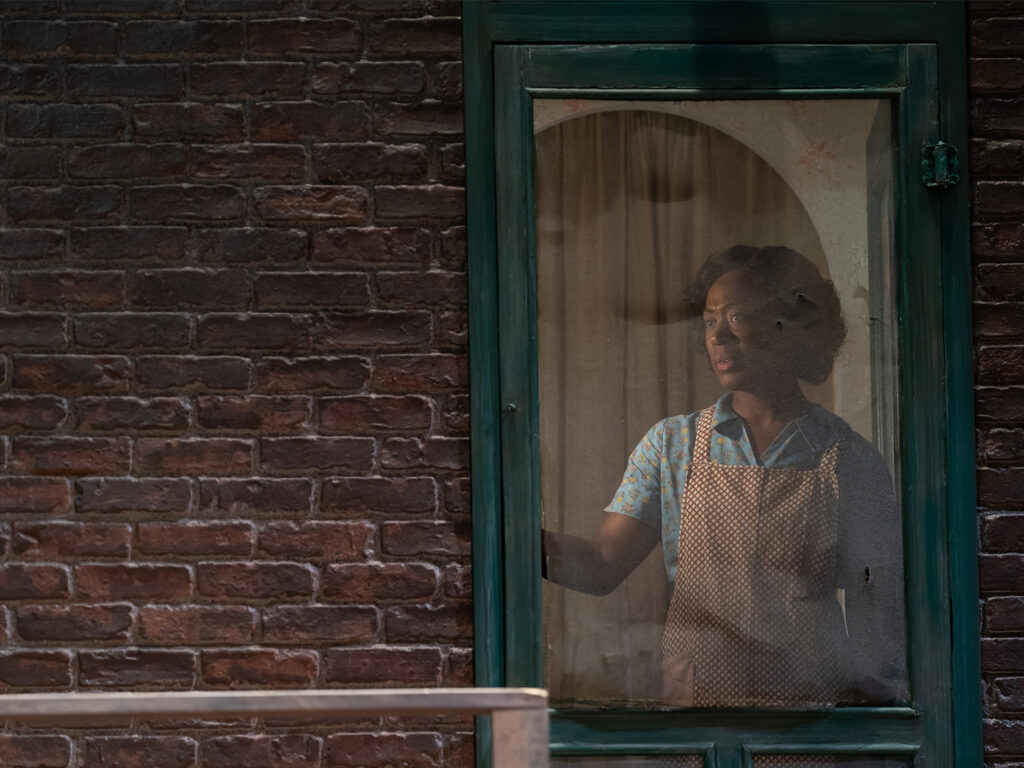 A woman stands inside the screen door of her 1950s row home, peering out into the yard. She wears a short-sleeved blue dress covered by a brown and white apron.