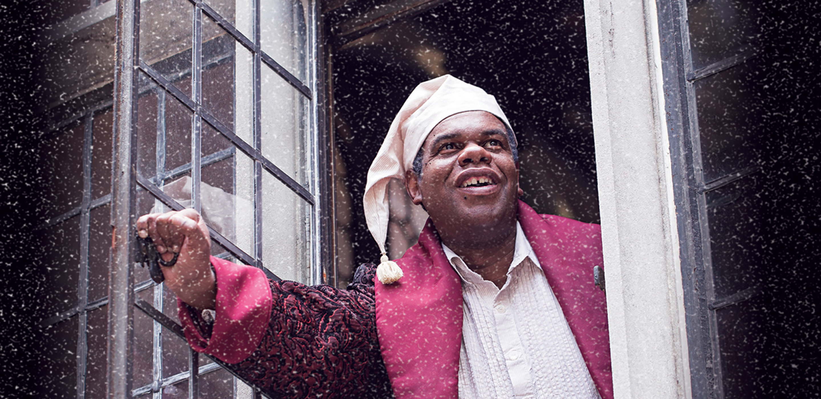 A smiling man dressed in Victorian era nightclothes opens a window to look out at the street. Snow is falling.