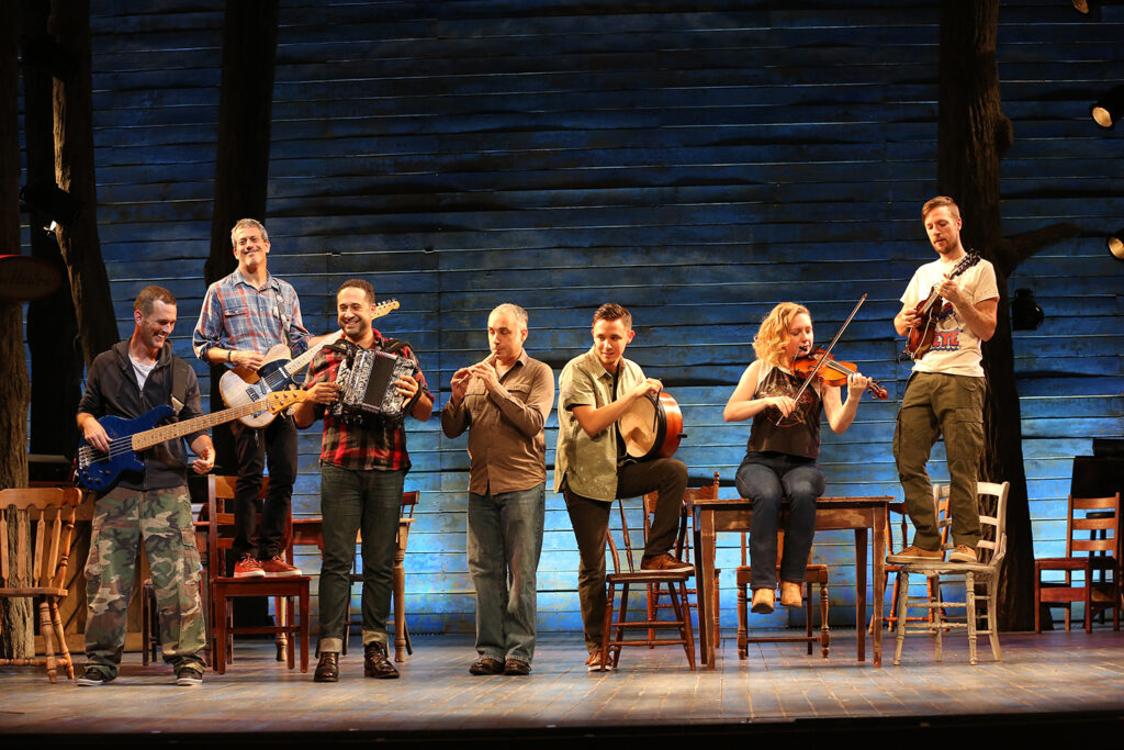 A group of seven musicians play a variety of instruments while standing on a stage set which resembles a wooden building. Some stand on chairs or sit on tables.