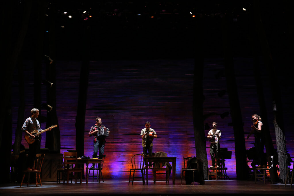 Five musicians stand on chairs and play instruments on a darkened stage.