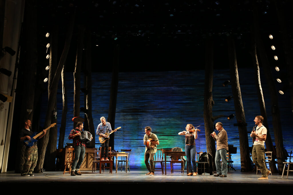A group of musicians play a variety of instruments on a stage set resembling a wooden building. They all look at a guitarist who stands on a chair.