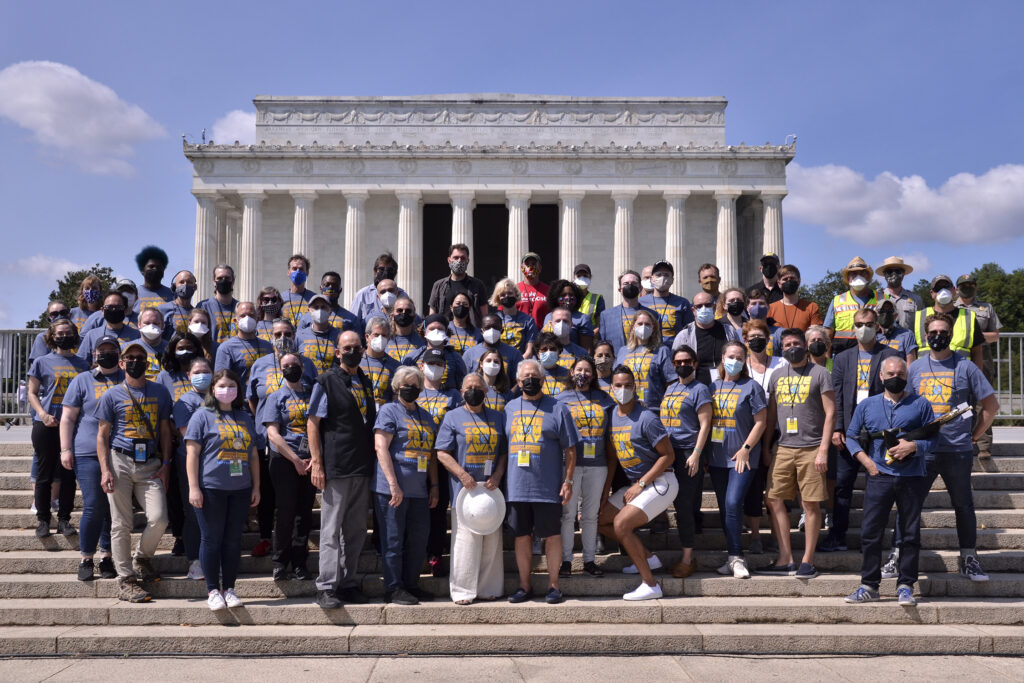 Seventy people stand on the lower steps of the Lincoln Memorial, wearing face masks and Come From Away t-shirts for the concert.