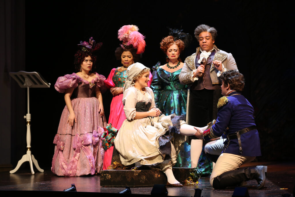 Cinderella is seated on a small stool as Cinderella’s Prince kneels to fits her golden shoe on her foot. Behind them, Cinderella’s Father, Stepmother and two sisters watch with varying expressions of disbelief, anger and opportunism.