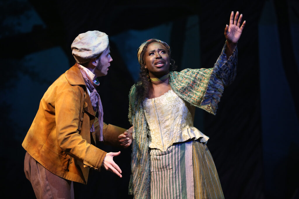 The Baker, wearing a yellow waistcoat, brown knee-length trousers and socks, green shoes and floppy baker’s hat, stands next to an actress playing his wife. She wears a corseted top, a brightly patterned scarf of reds, blues and yellows on her head, and an ankle-length yellow skirt. He stands bewildered with mouth open while she looks to the skies with her hand extended
