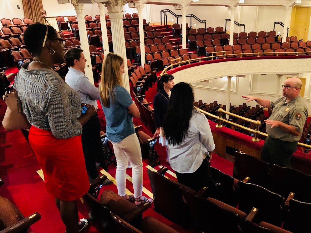 A group of young people stand on the upper tier of seating in Ford's Theatre. They watch a National Park Service ranger has he gives a tour of the theatre.