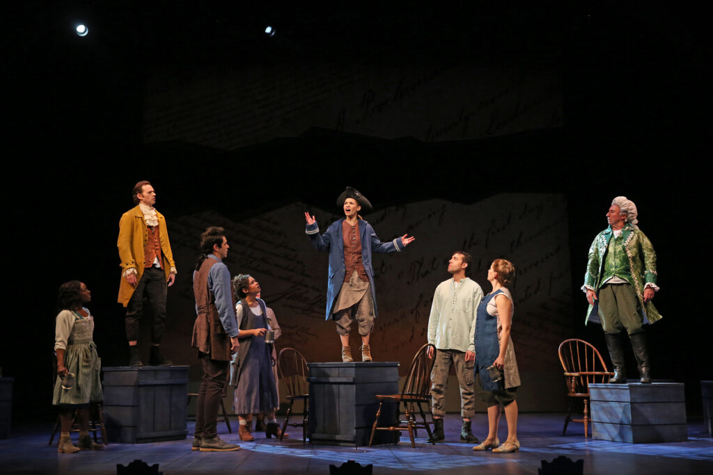 At center is an actress dressed as Patrick Henry. She stands on a large crate. A crowd gathers around her to listen to her words. They stand in front of a larger-than-life projection of the words from the Declaration of Independence.