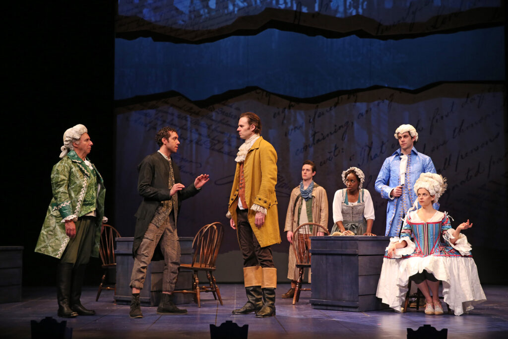James Hemmings, an enslaved man, addresses Thomas Jefferson while Ben Franklin listens. Also listening are a young man, an enslaved servant and a man and woman dressed in ostentatious 18th-century-style clothing and wigs made of paper.