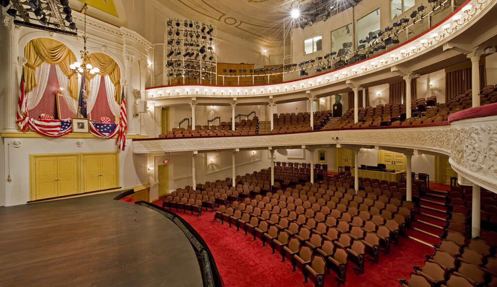 A side view of the stage and seating at Ford’s Theatre. On the left is the Presidential Box with an American flag, a framed picture of George Washington and American flag bunting draped over the box.