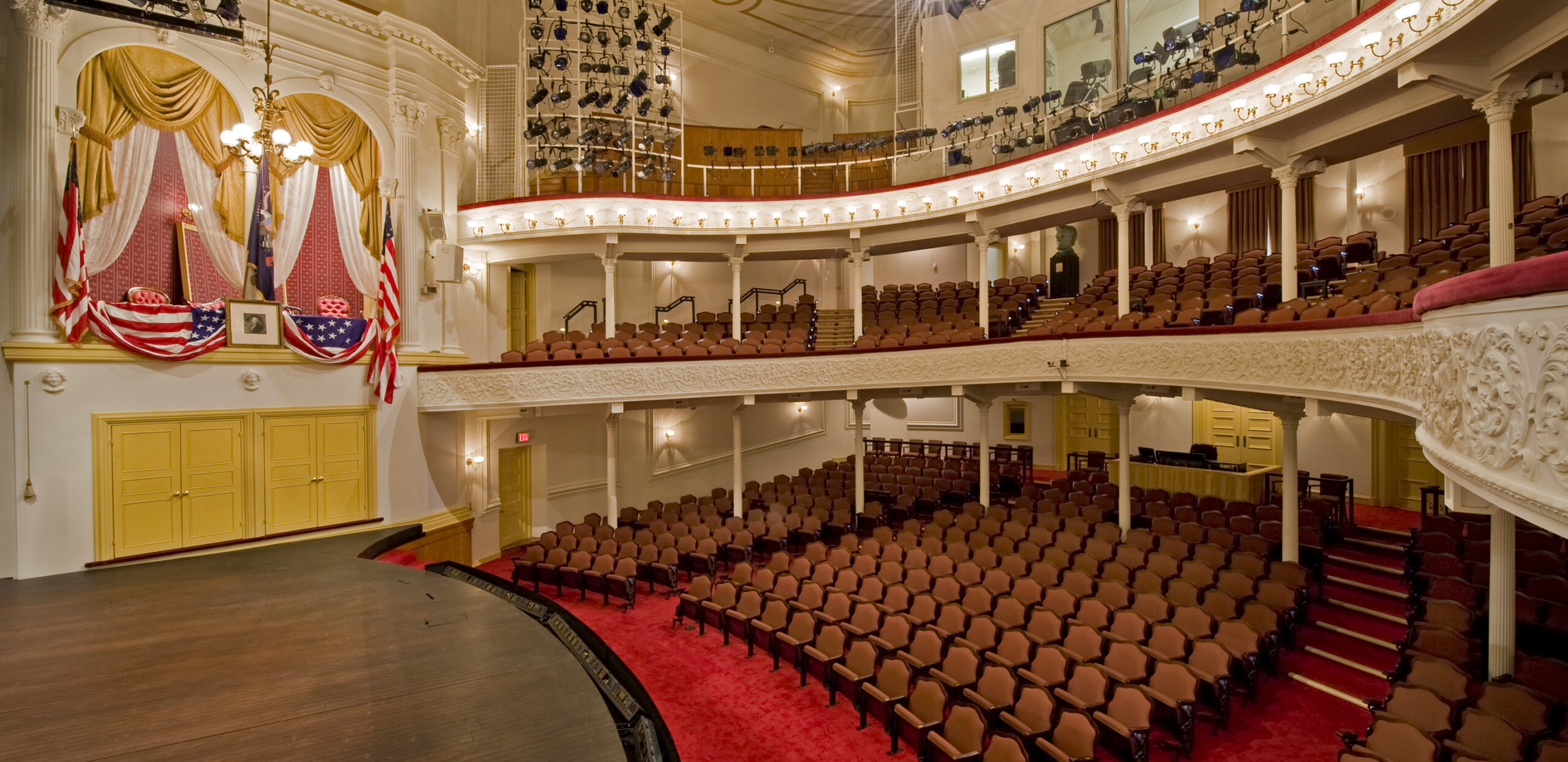 A side view of the stage and seating at Ford’s Theatre. On the left is the Presidential Box with an American flag, a framed picture of George Washington and American flag bunting draped over the box.