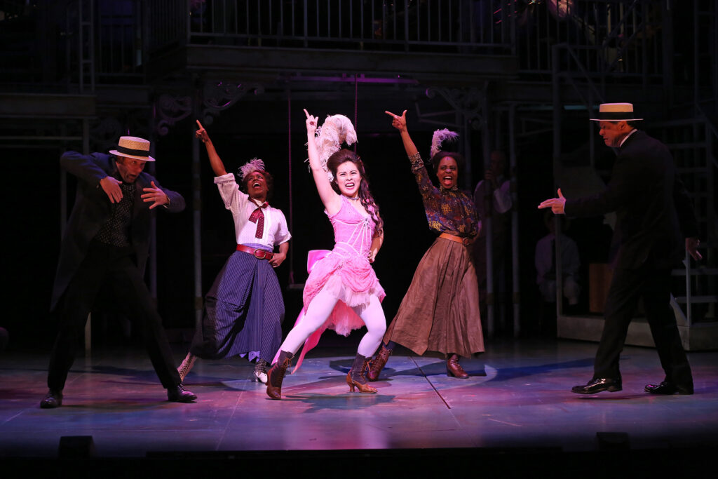 Three women dressed in burlesque outfits dance together while two men in suits and straw hats look on.