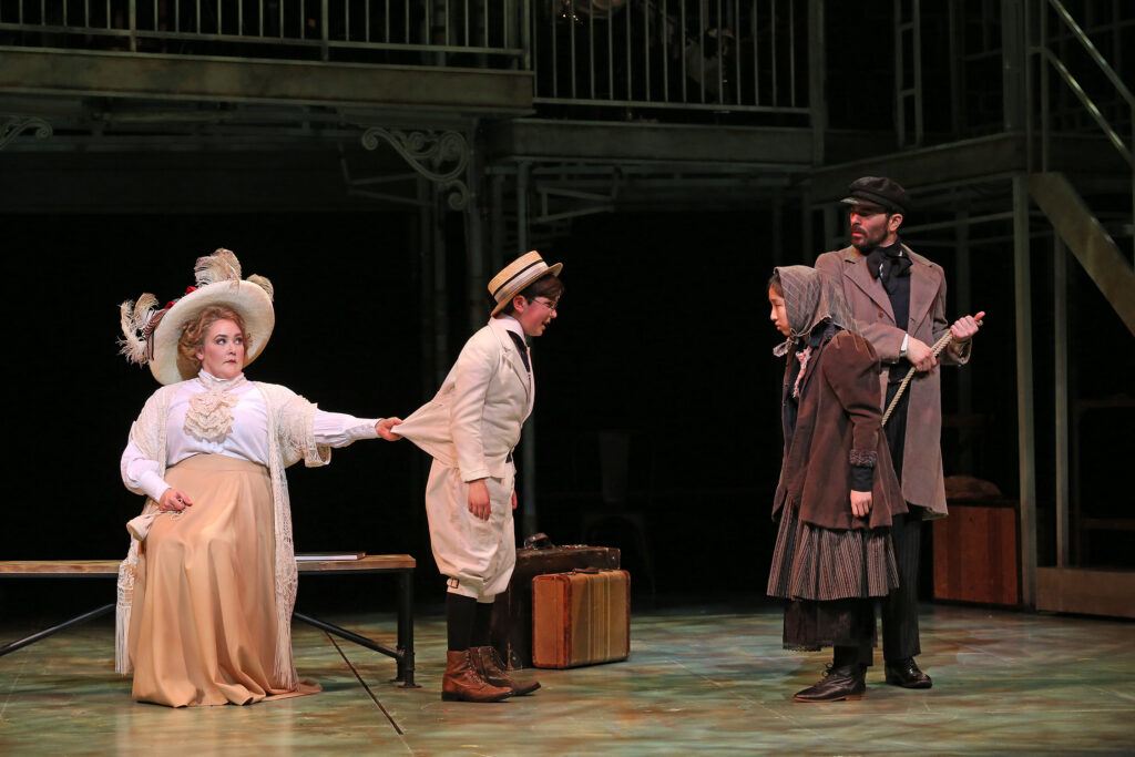 A young boy in a suit and straw hat talks to a young girl in ragged clothes. A woman sits on a bench and pulls on the back of the boy's suit jacket, while a man stands behind the young girl and looks at the boy suspiciously.