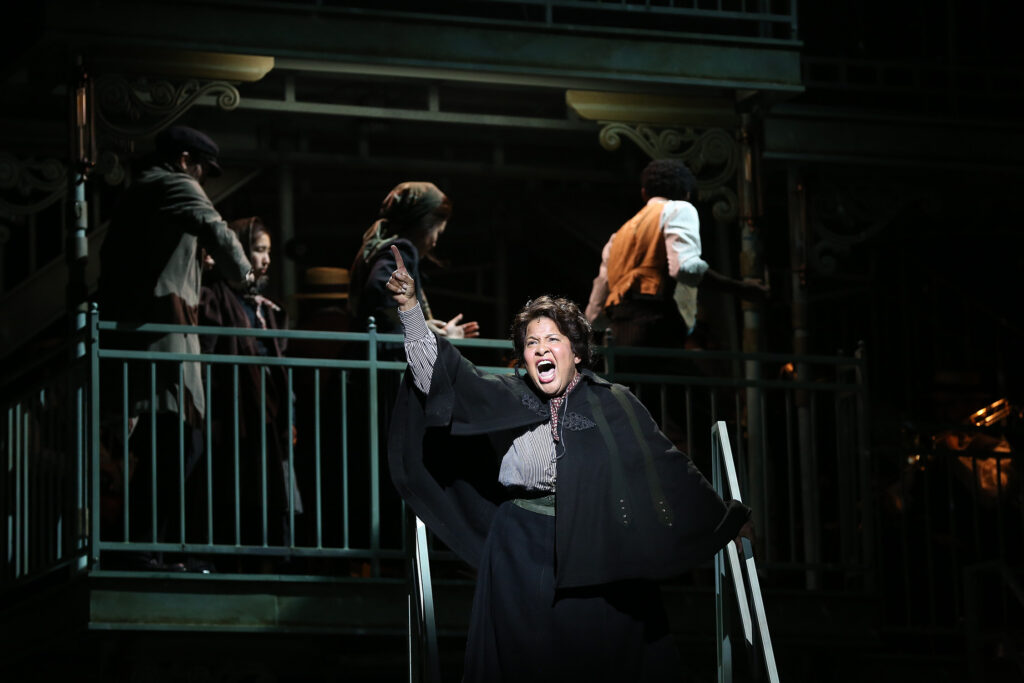 A woman in a black shawl stands on a staircase and sings. Behind her people wait in line on a balcony.