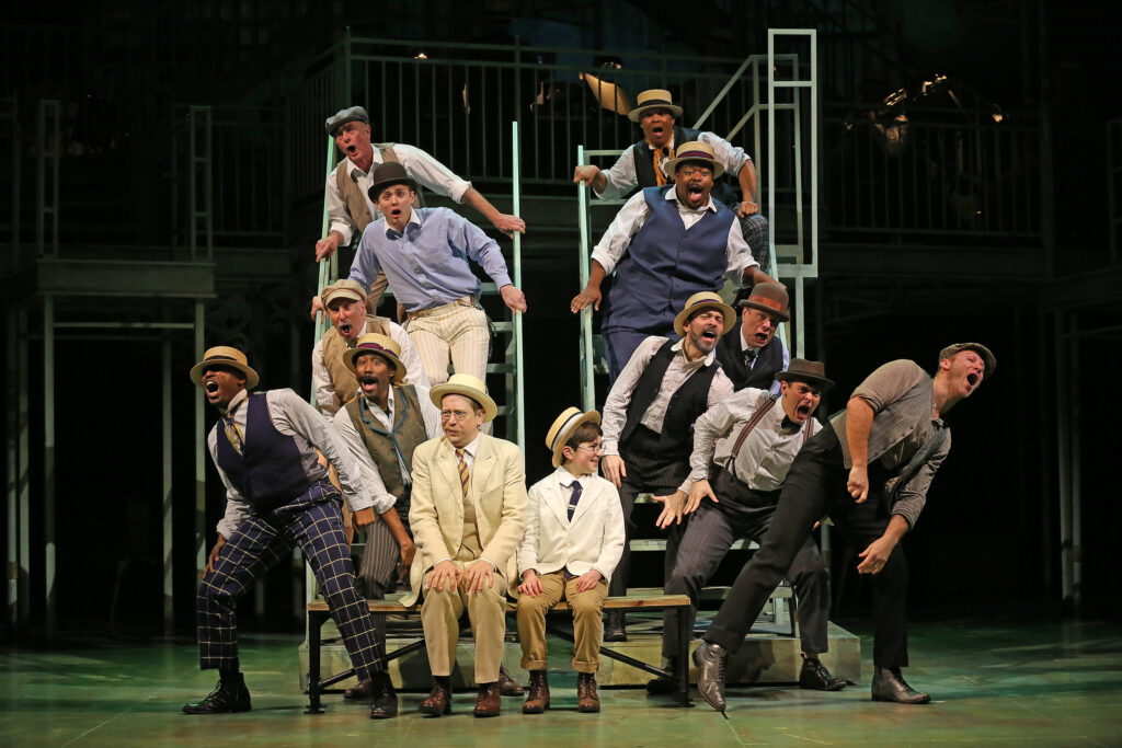 A man and young boy sit on a bench while people sing all around them. All are dressed in turn of the century outfits.