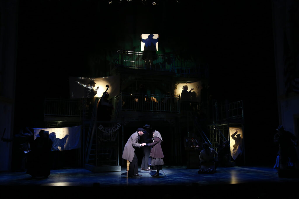 A man holds the hands of a young girl and talks to her. In the background, peoples shadows are projected against sheets all over a stage.