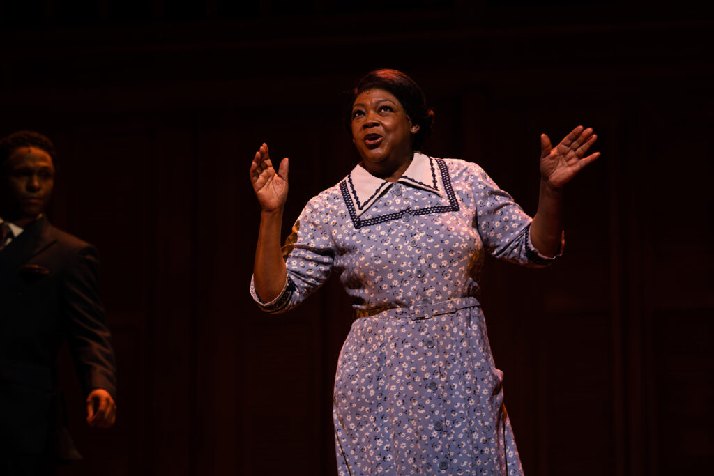 An African American woman stands center stage with her arms slightly up, wearing a blue dress with white flowers. In the background, an African American man wearing a black suit looks on.