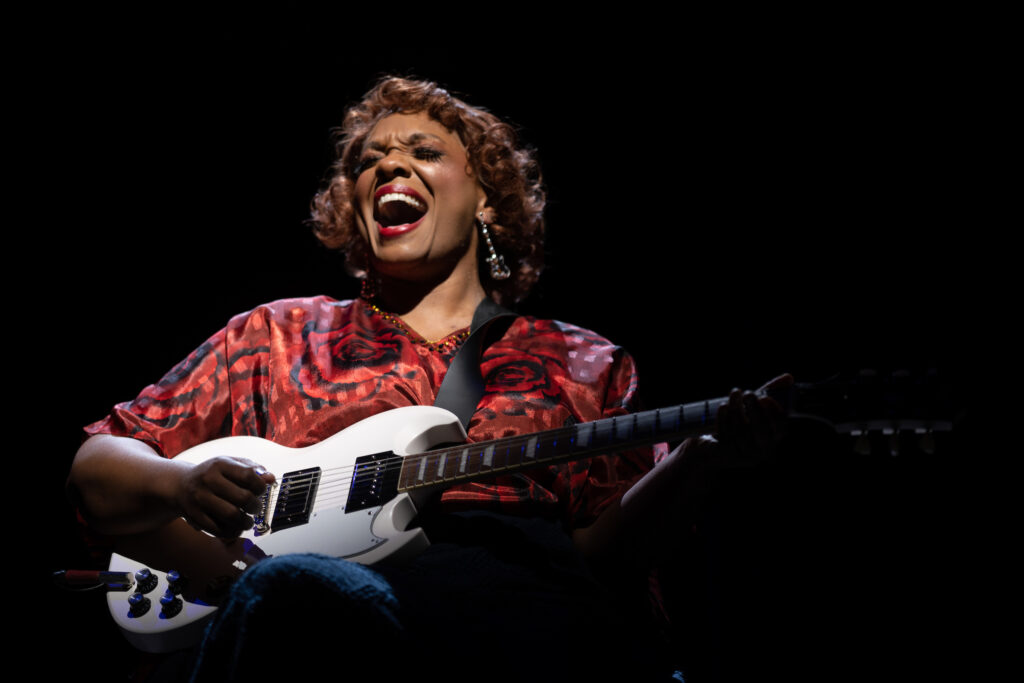 An African American woman wearing a red dress sits in a wheelchair center stage holding and playing a white electric guitar, singing with expression.