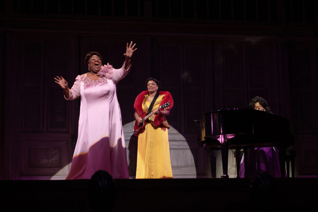 An African American woman wearing a wedding dress stands center stage on a trellis and plays an acoustic guitar, as the ensemble of wedding guests flanks her to her right and left. Circular lights and an onstage band are visible on a platform above her.