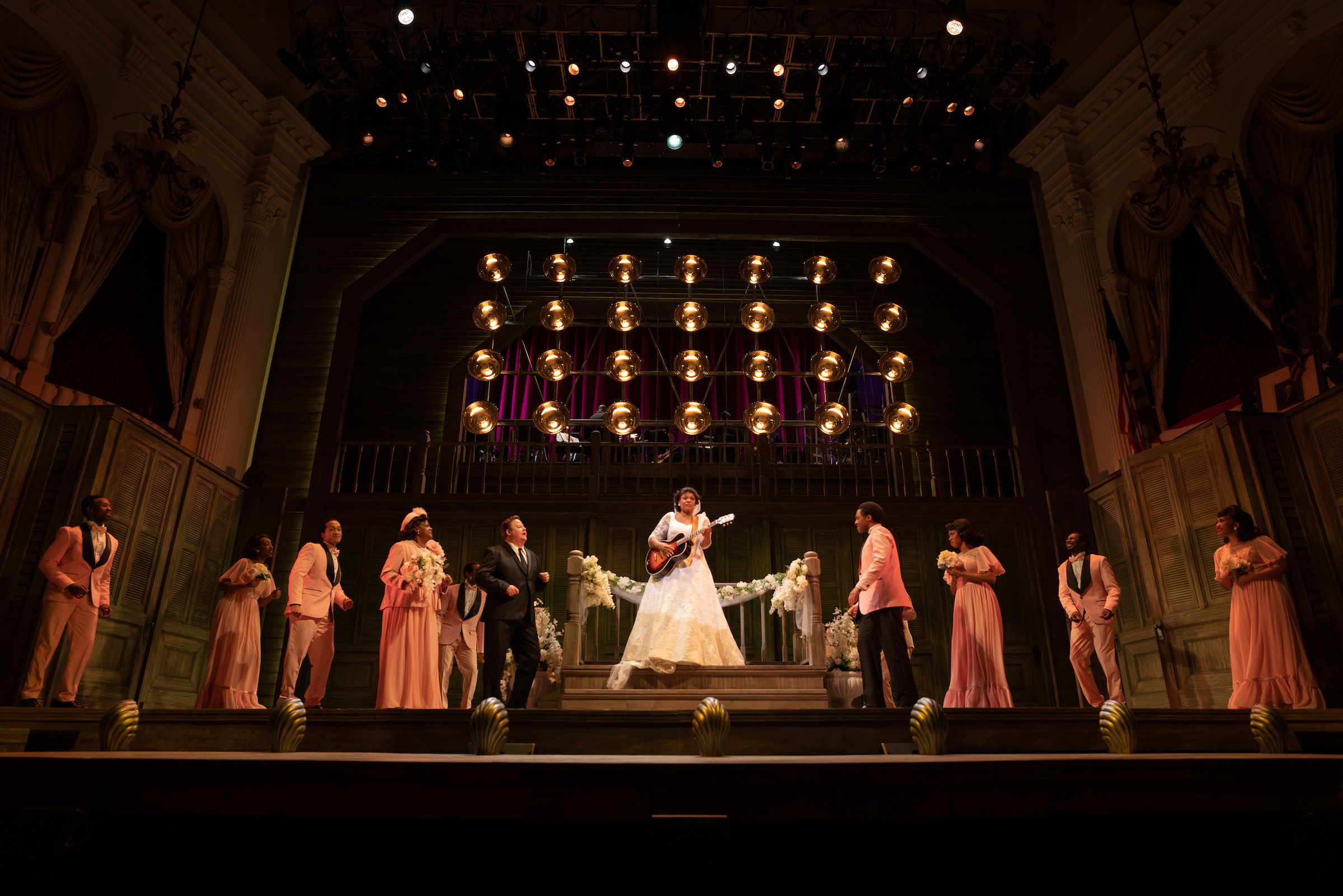 An African American woman wearing a wedding dress stands center stage on a trellis and plays an acoustic guitar, as the ensemble of wedding guests flanks her to her right and left. Circular lights and an onstage band are visible on a platform above her.