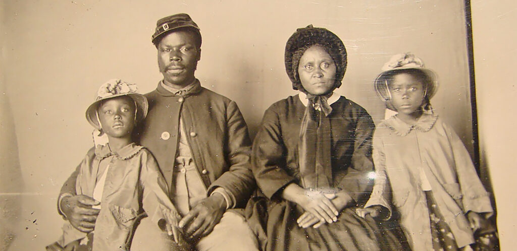 Black and white photograph from between 1963 and 1865, of an African American man and woman and their two daughters. The man is in Union uniform, his wife wears a fashionable dress and bonnet with satin ribbons, their daughters are on either side of the c