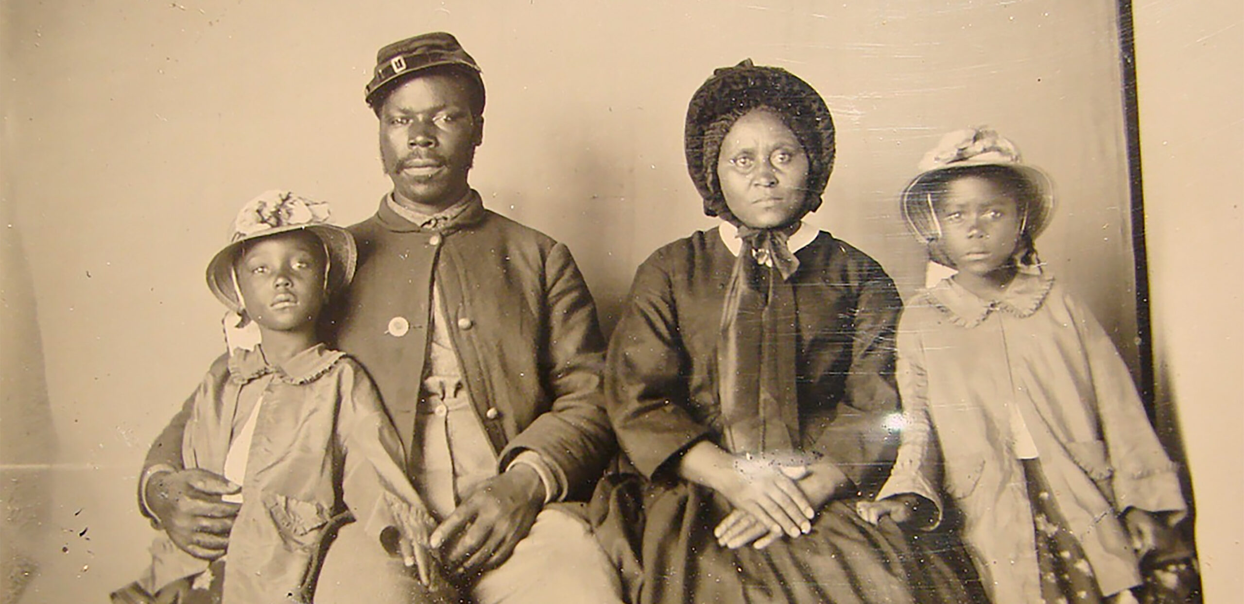 Black and white photograph from between 1963 and 1865, of an African American man and woman and their two daughters. The man is in Union uniform, his wife wears a fashionable dress and bonnet with satin ribbons, their daughters are on either side of the c