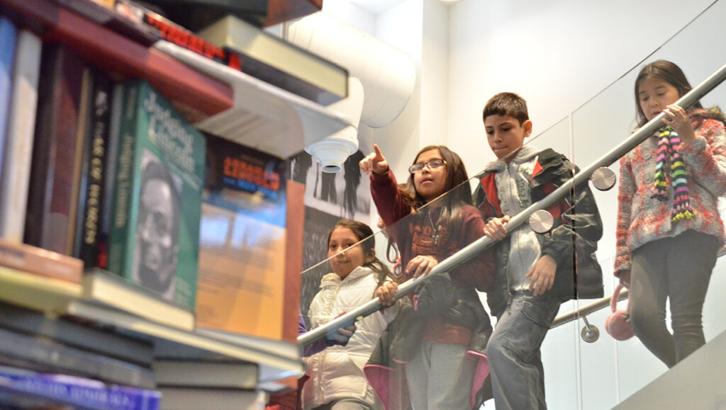 Four young children stand on a staircase looking at a large tower composed of books about Abraham Lincoln. A young girl points to the tower.