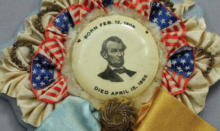 A mourning ribbon for President Lincoln with his portrait, birth and death dates in the center. Surrounding the portrait are small American flags. Hanging from the bottom are blue and hello ribbons and two decorative tassels.