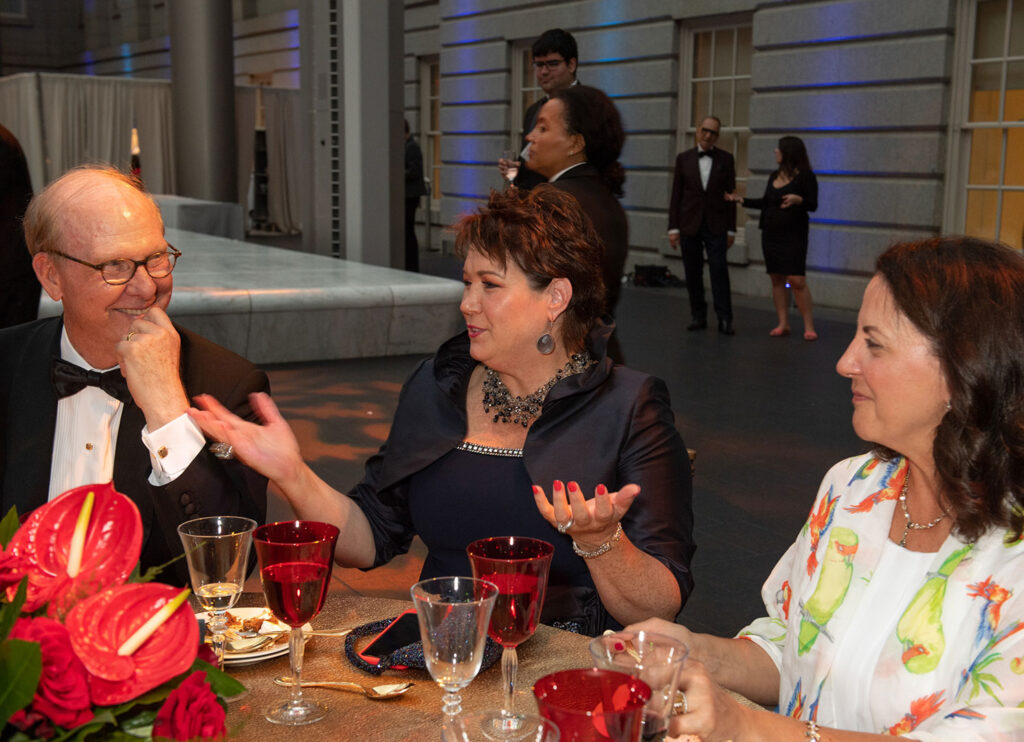 Two women and a man all chat together at a formal event while sitting at a table.