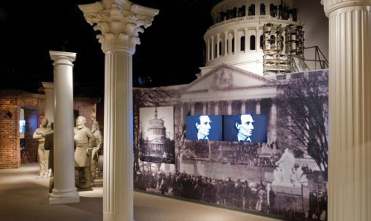 Inside the Ford’s Theatre Museum, two television screens display close-up images of Abraham Lincoln’s face. The screens are embedded in a display that is a large-scale photograph of the partially constructed U.S. Capitol dome taken during the Civil War. In front of the display are three white columns, designed to evoke the Capitol. Behind the display is a model of the partially constructed dome.