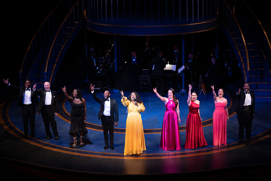 A group of men and women dressed in formal attire stand on a stage and sing. Behind them are two staircases leading up to a balcony. Above them lights are suspended from wires.