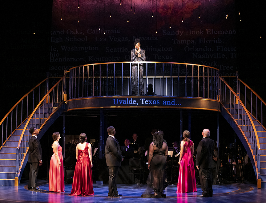A woman singing into a microphone on a stage balcony looks down at 7 performers.