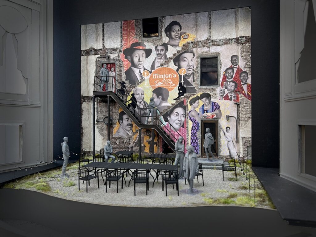 Set model of a grey-brick row house painted with a three-story wall mural. Mural features the faces of historic Black Philadelphian caterers and chefs. A series of metal fire-escape stairways lead to the three levels of the row house. In the courtyard below are arranged several rectangular tables and chairs.