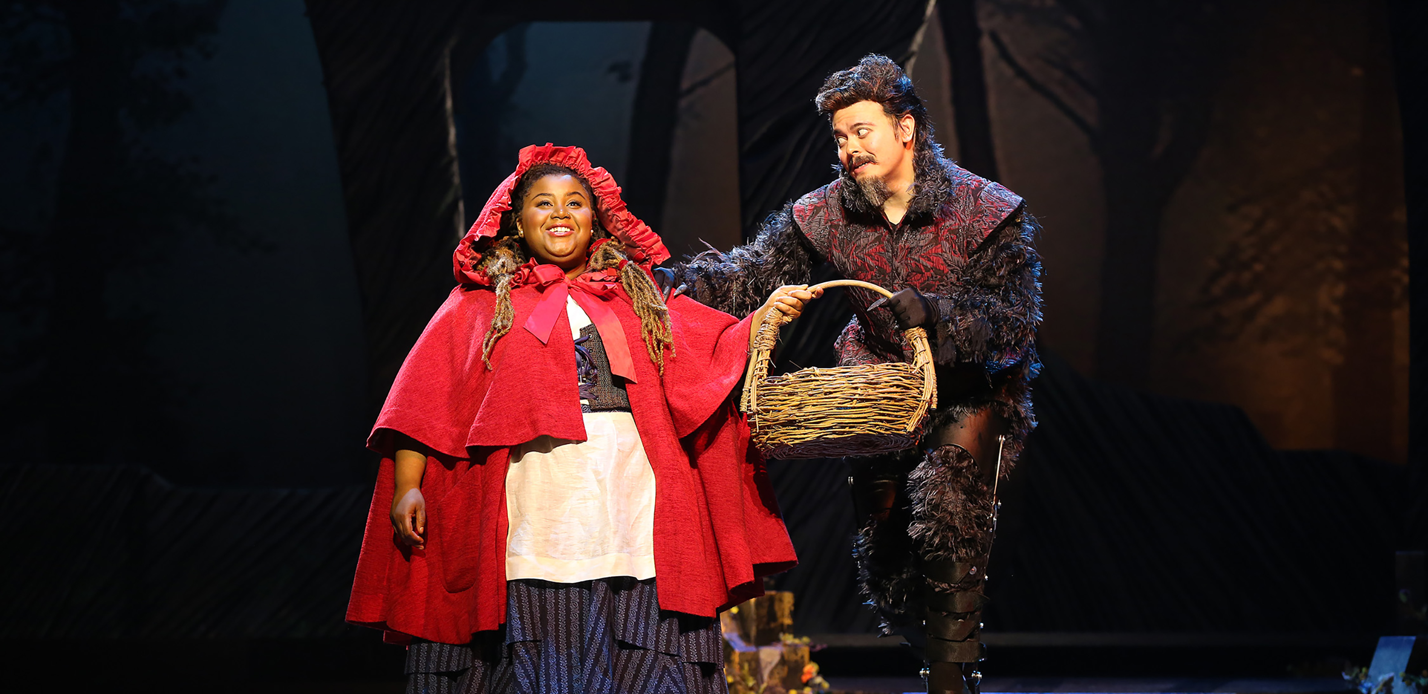 Little Red Ridinghood stands carrying a large wicker basket. On the right is an actor dressed as a wolf with one hand on her shoulder. His costume includes black stilts on his legs
